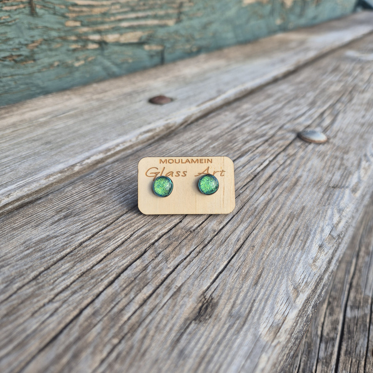 A pair of green textured glass stud earrings mounted on a small wooden display card engraved with 'MOULAMEIN Glass Art', placed on a weathered wooden surface.